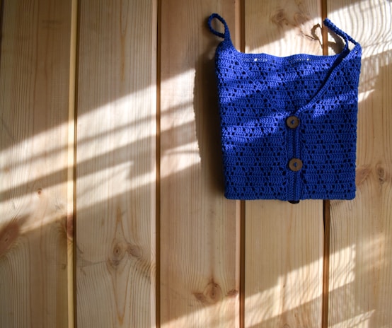 A bright blue crocheted bag with two wooden buttons is hanging against a light wooden wall. Sunlight filters in, casting shadows and highlighting the texture of the crochet work.