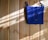 A bright blue crocheted bag with two wooden buttons is hanging against a light wooden wall. Sunlight filters in, casting shadows and highlighting the texture of the crochet work.