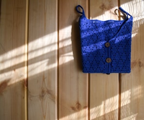 A bright blue crocheted bag with two wooden buttons is hanging against a light wooden wall. Sunlight filters in, casting shadows and highlighting the texture of the crochet work.