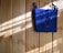 A bright blue crocheted bag with two wooden buttons is hanging against a light wooden wall. Sunlight filters in, casting shadows and highlighting the texture of the crochet work.