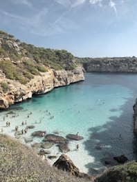 A serene cove with crystal clear turquoise water surrounded by rocky cliffs and lush greenery. Numerous people are enjoying the shallow waters and sandy beach area. The sky is partly cloudy, casting a soft light over the scene.