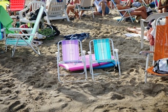 Several colorful beach chairs are arranged on sandy ground, surrounded by people relaxing. Towels and personal items are scattered around, indicating a busy beach scene. Various people in swimwear are sitting and lounging among the chairs.