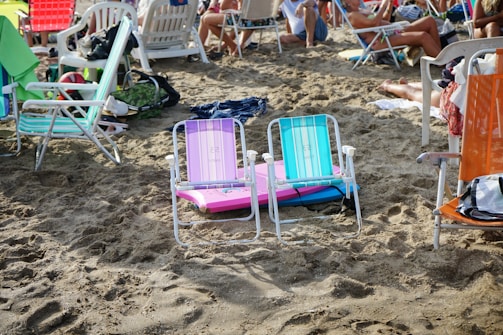Several colorful beach chairs are arranged on sandy ground, surrounded by people relaxing. Towels and personal items are scattered around, indicating a busy beach scene. Various people in swimwear are sitting and lounging among the chairs.