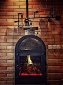 A rustic-looking metal stove is set against a backdrop of brown brickwork walls, featuring visible piping and a gauge. The stove has a glass door through which a warm glow is emitted, suggesting a fire burning inside.