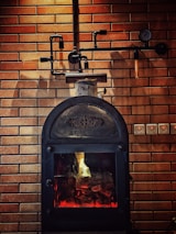 A rustic-looking metal stove is set against a backdrop of brown brickwork walls, featuring visible piping and a gauge. The stove has a glass door through which a warm glow is emitted, suggesting a fire burning inside.
