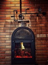 A rustic-looking metal stove is set against a backdrop of brown brickwork walls, featuring visible piping and a gauge. The stove has a glass door through which a warm glow is emitted, suggesting a fire burning inside.