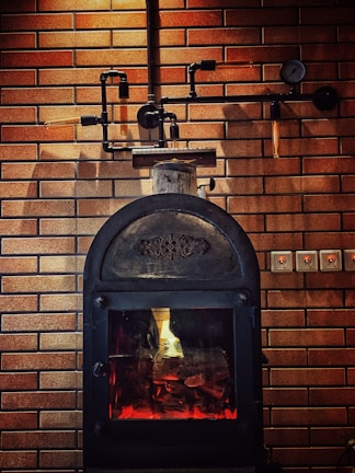 A rustic-looking metal stove is set against a backdrop of brown brickwork walls, featuring visible piping and a gauge. The stove has a glass door through which a warm glow is emitted, suggesting a fire burning inside.