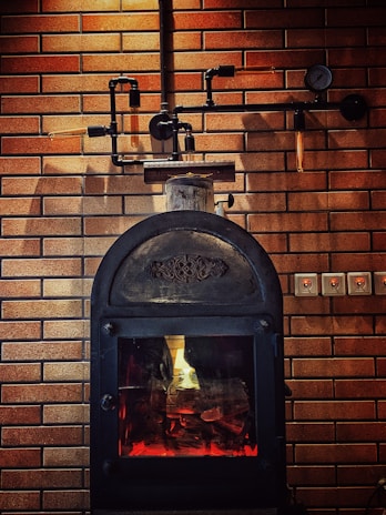 A rustic-looking metal stove is set against a backdrop of brown brickwork walls, featuring visible piping and a gauge. The stove has a glass door through which a warm glow is emitted, suggesting a fire burning inside.