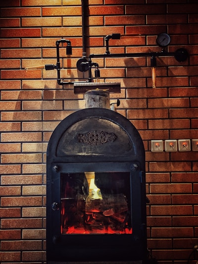 A rustic-looking metal stove is set against a backdrop of brown brickwork walls, featuring visible piping and a gauge. The stove has a glass door through which a warm glow is emitted, suggesting a fire burning inside.