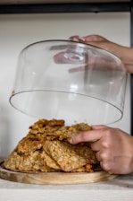 A person gently lifts a glass dome cover from a wooden plate full of freshly baked cookies, which are stacked on top of each other. The cookies appear to be oatmeal or similar, with visible chunks of nuts or chocolate. The setting is minimalistic, with a light background that highlights the warm tones of the cookies.