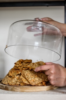 A person gently lifts a glass dome cover from a wooden plate full of freshly baked cookies, which are stacked on top of each other. The cookies appear to be oatmeal or similar, with visible chunks of nuts or chocolate. The setting is minimalistic, with a light background that highlights the warm tones of the cookies.