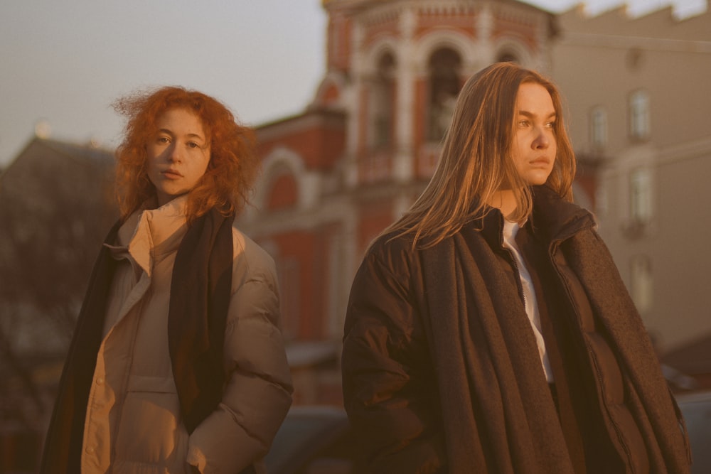 낮 동안 갈색 콘크리트 건물 근처에 서 있는 검은 코트를 입은 여성 2명