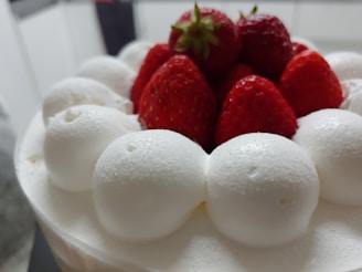 A cake topped with fresh red strawberries and dollops of white whipped cream. The surface of the cake appears smooth, and the strawberries are plump and glossy, suggesting freshness. The focus is on the cake with a blurred background, indicating depth.