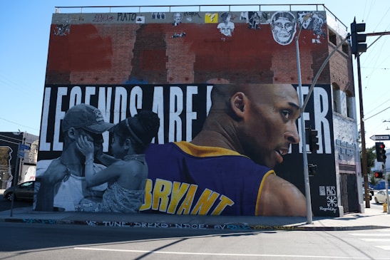 A large mural painted on the side of a brick building features an athlete in a basketball jersey with the name 'Bryant' visible. The background has large block letters with parts obscured by images. In the foreground, a couple is shown in black and white embracing closely with their foreheads touching.