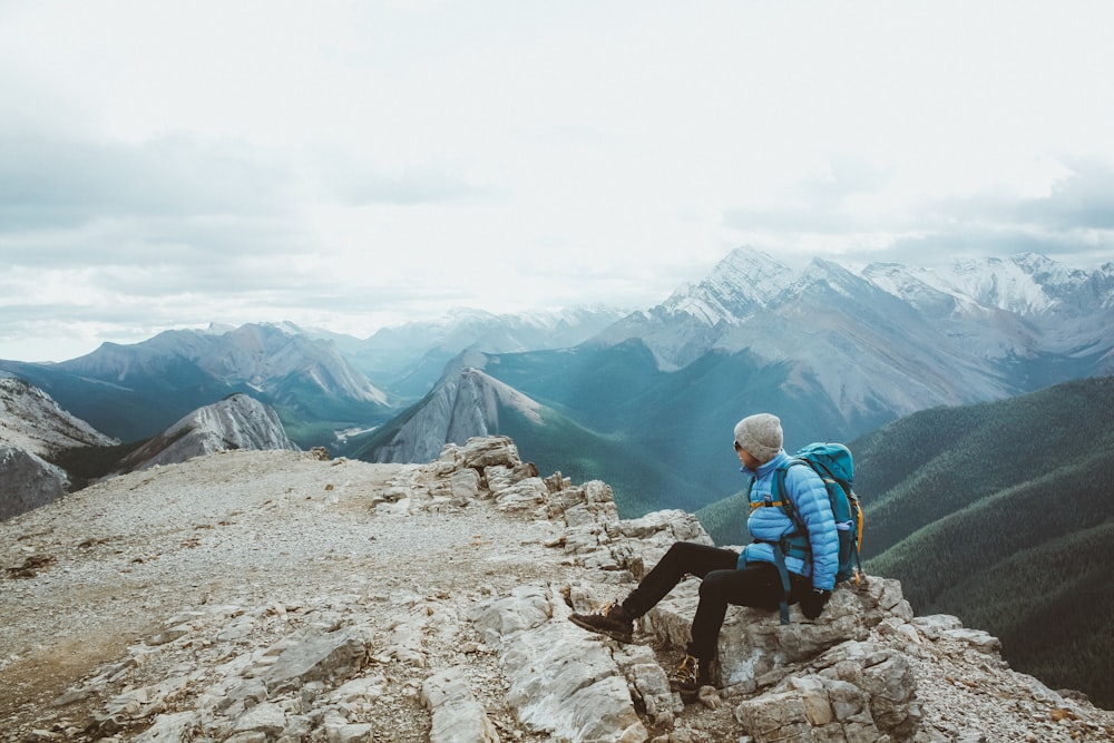 homme en veste noire assis sur un rocher près d’une montagne enneigée pendant la journée