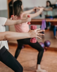 People are engaged in a fitness class, holding red dumbbells with both arms extended. The focus is on their arms and hands, with a blurred background showing a mirror and exercise balls. Participants are wearing athletic attire.