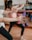 People are engaged in a fitness class, holding red dumbbells with both arms extended. The focus is on their arms and hands, with a blurred background showing a mirror and exercise balls. Participants are wearing athletic attire.