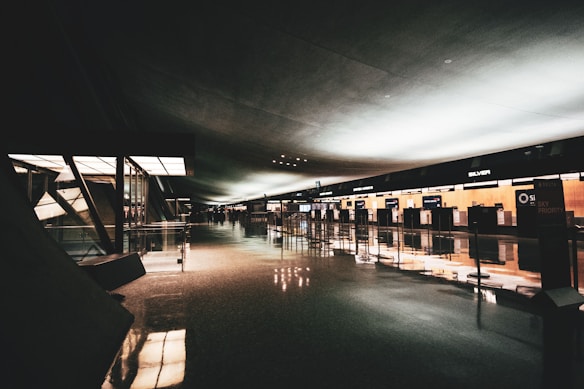 A spacious, dimly lit airport terminal with a modern design. The terminal features high ceilings and large glass windows along the left side. Reflective flooring stretches across the hall, and check-in counters line the right side, with signs for various airlines visible. The lighting creates a calm atmosphere.
