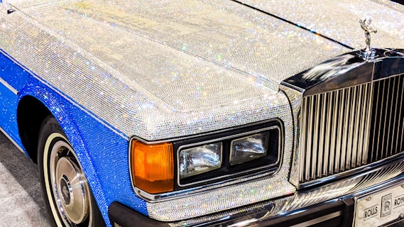 A luxury car adorned with sparkling crystals covering the hood and parts of the body. The car has a blue and silver color scheme with a distinctive grille and emblem on the front. The headlights are rectangular, and the vehicle exudes an opulent and extravagant appearance.