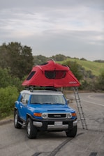 A blue SUV is parked on the side of a rural road with a red rooftop tent set up on the top. The tent is accessible by a ladder and is surrounded by greenery and a distant view of hills. The sky is overcast, adding a slightly moody atmosphere.