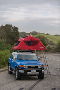 A blue SUV is parked on the side of a rural road with a red rooftop tent set up on the top. The tent is accessible by a ladder and is surrounded by greenery and a distant view of hills. The sky is overcast, adding a slightly moody atmosphere.
