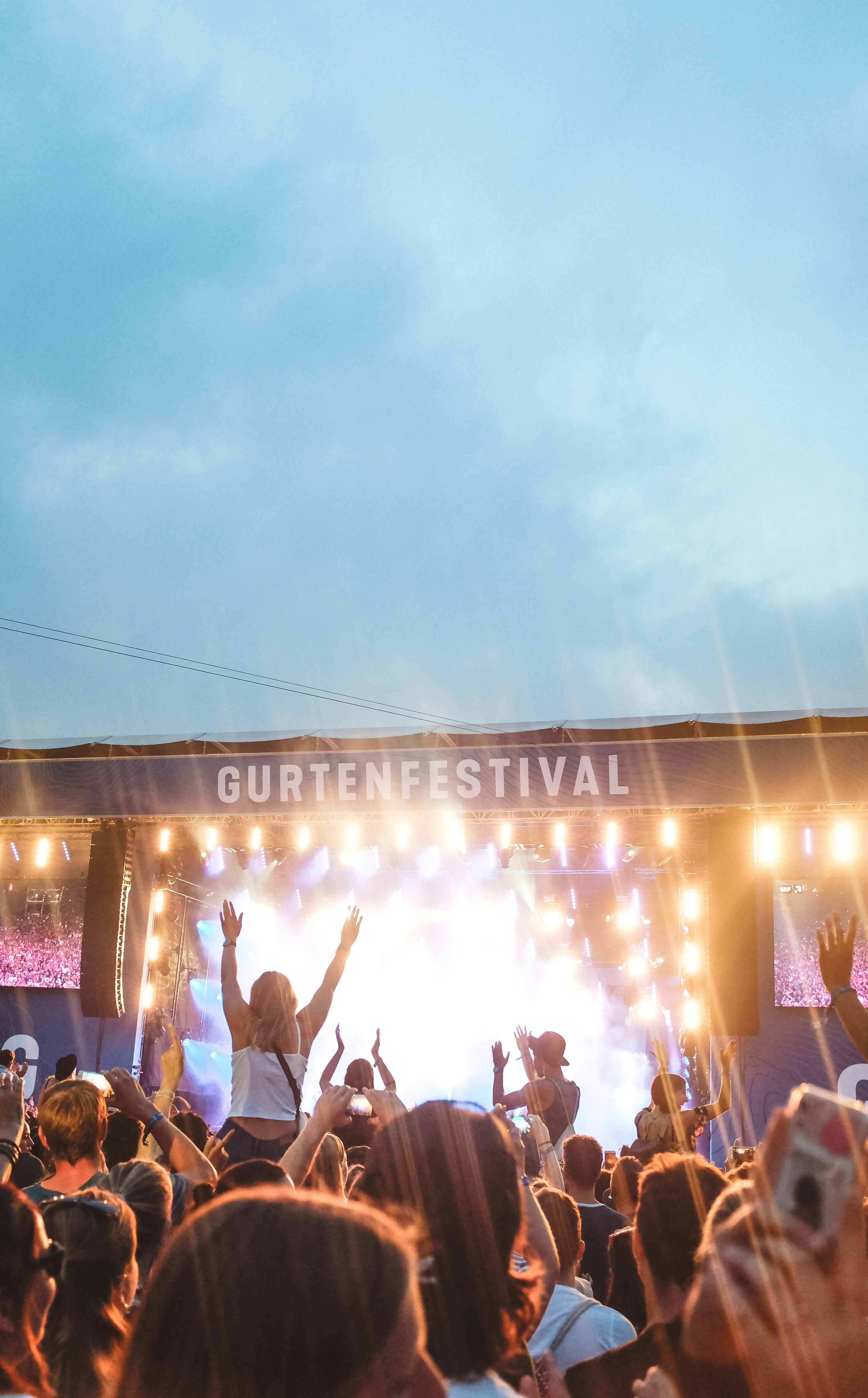 Festival crowd at dusk with colorful stage lighting, silhouettes of people raising hands, atmospheric outdoor concert setting