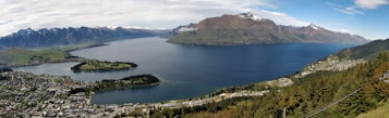 A panoramic view of a scenic landscape featuring a large lake surrounded by mountainous terrain. The foreground includes a town with clusters of buildings nestled among greenery, while the background reveals snow-capped peaks. The sky is partly cloudy, adding to the dramatic effect of the natural scenery.
