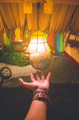 A hand reaching towards a decorative light fixture that is shaped like a globe. The fixture emits a warm glow and hangs from a ceiling of dried palm leaves. The background features a garden-like setting with greenery and colored walls.