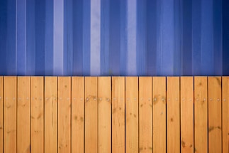 A vertical composition featuring a wooden fence with horizontal planks in the lower half, displaying a series of evenly spaced screws and a natural grain pattern. Above it, a corrugated metal wall in shades of blue presents vertical lines and slight weathering marks.