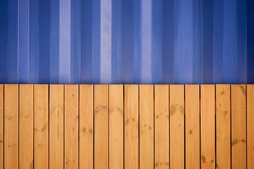 A vertical composition featuring a wooden fence with horizontal planks in the lower half, displaying a series of evenly spaced screws and a natural grain pattern. Above it, a corrugated metal wall in shades of blue presents vertical lines and slight weathering marks.