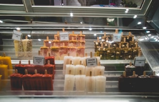 An assortment of artisanal popsicles are displayed in a glass showcase, each with a small sign indicating the flavor and price. The flavors include a variety of choices such as coco choco, strawberry cheesecake, watermelon raspberry, lemon, s'mores, and chocolate. The popsicles are arranged neatly in rows against a backdrop of a reflective glass surface, and there is lighting that highlights them.