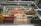 An assortment of artisanal popsicles are displayed in a glass showcase, each with a small sign indicating the flavor and price. The flavors include a variety of choices such as coco choco, strawberry cheesecake, watermelon raspberry, lemon, s'mores, and chocolate. The popsicles are arranged neatly in rows against a backdrop of a reflective glass surface, and there is lighting that highlights them.