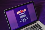 A laptop displays a screen with a vibrant purple background featuring the text 'Wash & Vax' with stylized virus iconography above it. Detailed text below outlines a call to action related to vaccines and social distancing, with a prominent 'start' button visible.