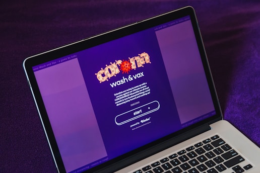 A laptop displays a screen with a vibrant purple background featuring the text 'Wash & Vax' with stylized virus iconography above it. Detailed text below outlines a call to action related to vaccines and social distancing, with a prominent 'start' button visible.