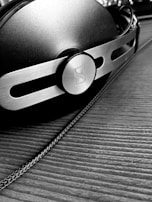 A close-up, black and white image of a pair of headphones with a metallic and modern design, featuring a prominent circular logo on the side. The surface is textured, and the headphones are resting on a wooden surface with a visible cable running alongside.