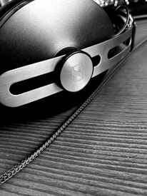 A close-up, black and white image of a pair of headphones with a metallic and modern design, featuring a prominent circular logo on the side. The surface is textured, and the headphones are resting on a wooden surface with a visible cable running alongside.