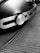 A close-up, black and white image of a pair of headphones with a metallic and modern design, featuring a prominent circular logo on the side. The surface is textured, and the headphones are resting on a wooden surface with a visible cable running alongside.