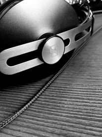 A close-up, black and white image of a pair of headphones with a metallic and modern design, featuring a prominent circular logo on the side. The surface is textured, and the headphones are resting on a wooden surface with a visible cable running alongside.