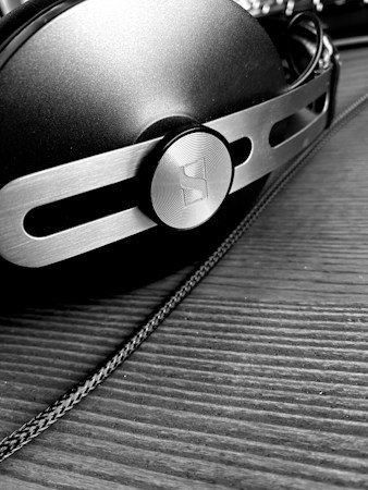 A close-up, black and white image of a pair of headphones with a metallic and modern design, featuring a prominent circular logo on the side. The surface is textured, and the headphones are resting on a wooden surface with a visible cable running alongside.