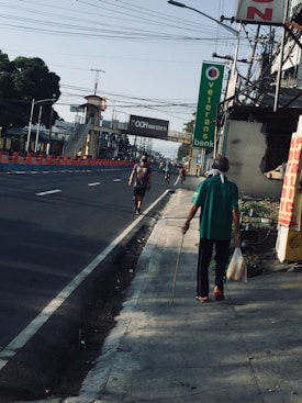 A street scene with an elderly person walking along a sidewalk, holding a stick and carrying plastic bags. The road is empty with a few people in the background, indicating early morning or a quiet time. Overhead wires stretch across the sky, and there&rsquo;s a row of orange and blue barricades on the left. A sign for Veterans Bank is visible on the right.