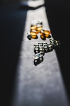 A collection of translucent capsules and pills scattered across a dark, textured surface. The lighting casts dramatic shadows and highlights, creating a contrast between the bright capsules and the dark background.