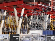 An intricate construction site with a complex framework of red steel beams and scaffolding stands prominently. Several white structural supports and yellow braces are visible, indicating an ongoing building project. The foreground displays signage in a language suggesting construction of luxury apartments.