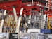 An intricate construction site with a complex framework of red steel beams and scaffolding stands prominently. Several white structural supports and yellow braces are visible, indicating an ongoing building project. The foreground displays signage in a language suggesting construction of luxury apartments.