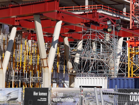 An intricate construction site with a complex framework of red steel beams and scaffolding stands prominently. Several white structural supports and yellow braces are visible, indicating an ongoing building project. The foreground displays signage in a language suggesting construction of luxury apartments.