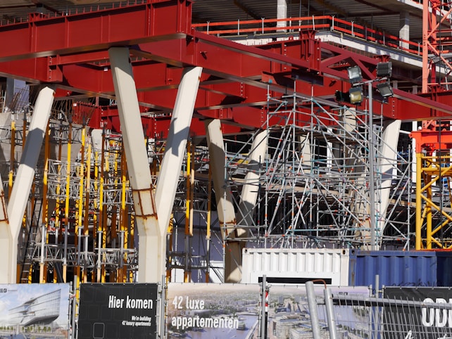 An intricate construction site with a complex framework of red steel beams and scaffolding stands prominently. Several white structural supports and yellow braces are visible, indicating an ongoing building project. The foreground displays signage in a language suggesting construction of luxury apartments.