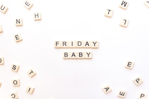 Scrabble tiles are arranged to spell out 'Friday Baby' on a white surface. Additional scattered tiles are visible around the main arrangement, creating a playful and casual atmosphere.