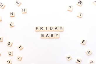 Scrabble tiles are arranged to spell out 'Friday Baby' on a white surface. Additional scattered tiles are visible around the main arrangement, creating a playful and casual atmosphere.