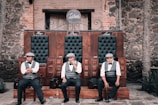 Three men in vests, bow ties, and hats seated on a wooden bench with three sections, each labeled with names Hugo, Paco, and Luis. The background consists of rustic brick and stone walls, and there is a wooden door behind them. The setting appears old-fashioned and reminiscent of early 20th-century establishments.