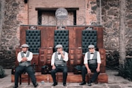 Three men in vests, bow ties, and hats seated on a wooden bench with three sections, each labeled with names Hugo, Paco, and Luis. The background consists of rustic brick and stone walls, and there is a wooden door behind them. The setting appears old-fashioned and reminiscent of early 20th-century establishments.