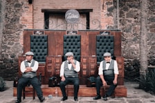 Three men in vests, bow ties, and hats seated on a wooden bench with three sections, each labeled with names Hugo, Paco, and Luis. The background consists of rustic brick and stone walls, and there is a wooden door behind them. The setting appears old-fashioned and reminiscent of early 20th-century establishments.